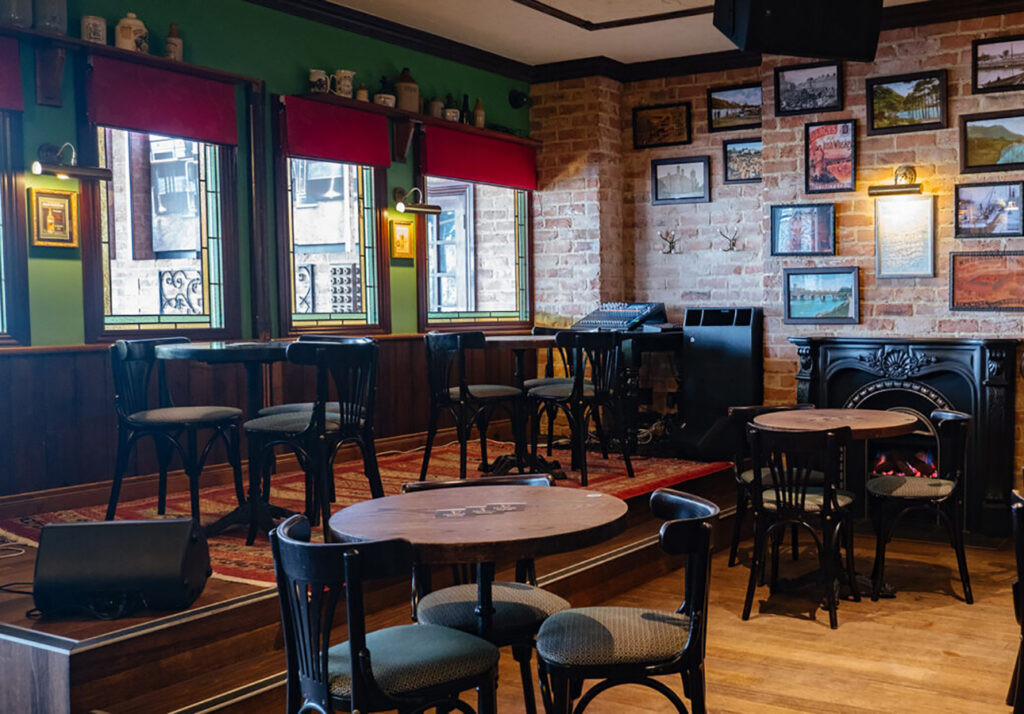 Seating area with small stage and fireplace at The Galway Hooker Irish Pub in Scarborough, Western Australia, featuring exposed brick walls, framed artwork, and timber finishes.