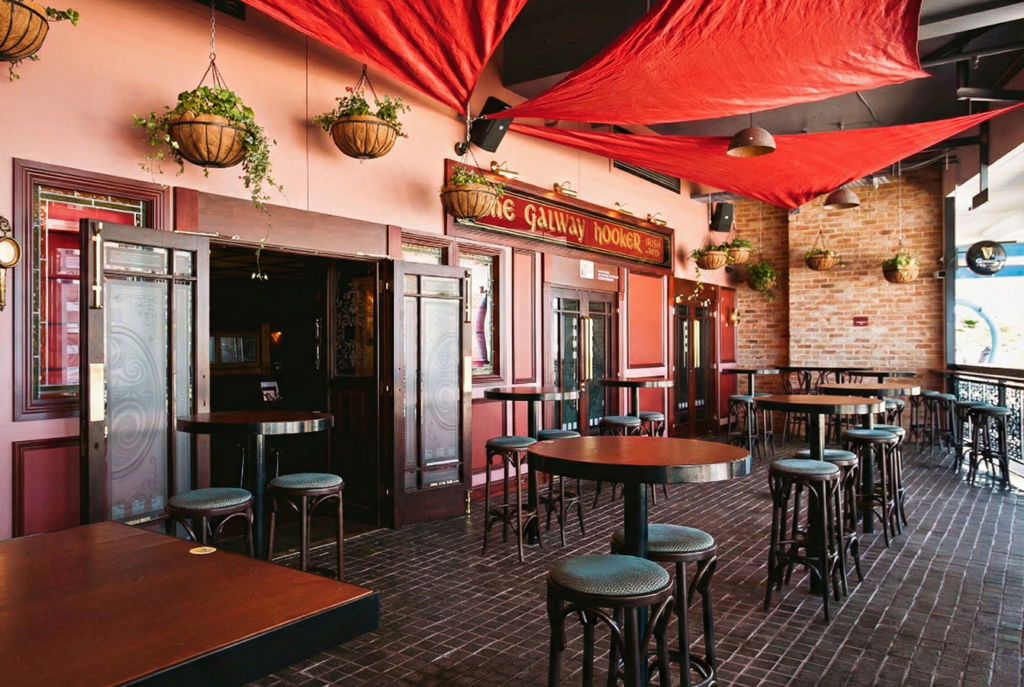 Covered terrace seating at The Galway Hooker Irish Pub in Scarborough, Western Australia, with high tables, brick walls, and red fabric ceiling canopies.