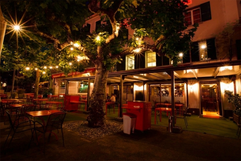 Night exterior and terrace seating of Red Piper gastropub in Vevey, Switzerland, with outdoor tables, tree lighting, and illuminated entrance.